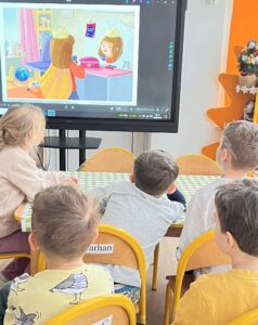 Na zdjęciu widać grupę małych dzieci siedzących przy stole w sali przedszkolnej. Dzieci siedzą na żółtych krzesełkach i patrzą na duży ekran, na którym wyświetlony jest obrazek przedstawiający dziewczynkę w koronie, przeglądającą się w lustrze. Scena na ekranie wygląda jak ilustracja z bajki lub książki dla dzieci.

W pomieszczeniu panuje ciepła, przyjazna atmosfera. W tle widać pomarańczowy regał w kształcie gwiazdy oraz ozdobioną choinkę lub stroik. Stół przykryty jest zielono-białym obrusem w kratkę. Dzieci wydają się skupione i zainteresowane tym, co oglądają.