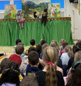 Na zdjęciu widać grupę dzieci siedzących na podłodze i oglądających przedstawienie kukiełkowe. Scena jest udekorowana zieloną zasłoną z falbaną, a w tle znajduje się kolorowa, malowana sceneria przedstawiająca górski krajobraz z lasem, drogą i górami.

Na scenie występują trzy kukiełki: po lewej stronie słonik w różowej spódniczce i białej bluzce, w środku czarno-biała sroka, a po prawej brązowy miś ubrany w kamizelkę i kapelusz. Po bokach sceny widać dodatkowe obrazy z bajkowymi zwierzętami – po lewej żółwia i biedronkę, a po prawej białego kota z pomarańczowym ogonem.

Dzieci siedzą w kilku rzędach, uważnie obserwując przedstawienie. Widać, że są skupione i zainteresowane, a cała scena ma radosny, kolorowy i przyjazny charakter, typowy dla spektakli teatralnych dla najmłodszych.