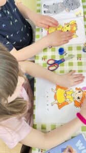 Na zdjęciu widać dwoje dzieci siedzących przy stole i wykonujących prace plastyczne. Na stole leży zielono-biały obrus w kratkę, a na nim rozłożone są kolorowanki przedstawiające bajkowe postacie – zwierzątka ubrane w kolorowe stroje. Dzieci kolorują obrazki kredkami lub flamastrami, a obok mają nożyczki, klej w sztyfcie i inne przybory plastyczne.

Dziecko po lewej stronie ma na sobie różową koszulkę, a dziecko po prawej granatową z jasnym wzorem. Atmosfera jest spokojna i twórcza – wygląda na to, że dzieci z zaangażowaniem pracują nad swoimi rysunkami.