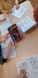 Na zdjęciu widać grupę dzieci siedzących przy prostokątnym stole i kolorujących obrazki w książeczkach do kolorowania. Na każdej stronie znajdują się postacie przypominające bajkowe zwierzątka lub leśne stworzonka ubrane w różne stroje.

Na środku stołu stoi metalowe pudełko wypełnione kredkami ołówkowymi i świecowymi w różnych kolorach. Dzieci mają przed sobą otwarte kolorowanki i skupiają się na swojej pracy. Jedno z dzieci ma na sobie żółtą bluzkę w białe wzory, inne brązową, a pozostałe ubrania w stonowanych kolorach. Scena przedstawia spokojny, twórczy moment zajęć plastycznych w przedszkolu lub wczesnoszkolnej sali.