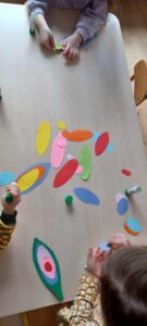 Na zdjęciu widać troje dzieci siedzących przy jasnym stole i wykonujących prace plastyczne. Na blacie rozłożone są kolorowe, owalne kawałki papieru w różnych rozmiarach i barwach — żółtych, czerwonych, różowych, zielonych, niebieskich i pomarańczowych. Dzieci trzymają w rękach kleje w sztyfcie i przyklejają elementy, tworząc z nich kompozycje przypominające wzory lub abstrakcyjne kształty.

Dziecko po lewej stronie ma na sobie bluzę w żółto-czarne paski, dziecko po prawej żółtą bluzę w białe wzory, a dziecko u góry zdjęcia jasnofioletową bluzę. Scena przedstawia moment wspólnej, kreatywnej zabawy plastycznej w przedszkolu lub wczesnoszkolnej sali.