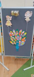 Na zdjęciu widać tablicę stojącą na białych nóżkach, ustawioną w sali przedszkolnej. Na ciemnoszarym tle tablicy przymocowane są kolorowe prace plastyczne wykonane przez dzieci.

Na dole znajduje się duży, niebieski paw z rozłożonym, efektownym ogonem złożonym z wielu barwnych piór w odcieniach zieleni, różu, pomarańczu, żółci i błękitu. Nad nim umieszczone są dwie postacie – dziewczynki z papieru, ubrane w kolorowe sukienki, przyklejone do patyczków, co sugeruje, że mogą być używane jako kukiełki. Między nimi, w górnej części tablicy, znajduje się złota korona z papieru.

Całość wygląda jak element przedstawienia lub zajęć teatralnych dla dzieci, prawdopodobnie związanych z bajką lub opowieścią o pawiu i królewskiej koronie.