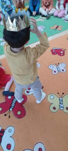 Na zdjęciu widać dziecko stojące na kolorowym dywanie w przedszkolu. Dywan jest ozdobiony dużymi motylami w różnych kolorach — czerwonym, żółtym, białym i zielonym. Dziecko ma na sobie jasnoszare spodnie, żółtą bluzkę i białe skarpetki. Na głowie ma złotą, błyszczącą koronę z papieru i trzyma ją obiema rękami, jakby poprawiało lub zakładało.

W tle, na zielonej części dywanu, siedzi grupa innych dzieci w kolorowych ubraniach, tworząc półkole. Scena wygląda na zabawę lub zajęcia tematyczne w przedszkolu, prawdopodobnie związane z królem, królową lub bajkową tematyką