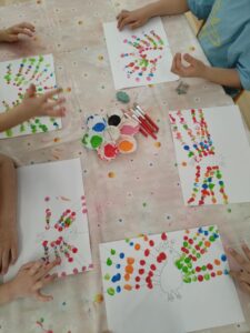 Na zdjęciu widać stół przykryty jasnym, lekko poplamionym obrusem, przy którym siedzi czworo dzieci. Na stole leżą kartki z narysowanymi konturami pawia, a dzieci ozdabiają jego ogon kolorowymi kropkami z farby. Używają do tego palców, tworząc barwne wzory w odcieniach czerwieni, zieleni, niebieskiego, pomarańczowego i różowego.

Na środku stołu stoi paleta z farbami w kilku kolorach oraz kilka pędzelków. Każde dziecko ma własną kartkę i pracuje nad swoim pawiem, dzięki czemu wszystkie prace różnią się układem i intensywnością kolorów. Scena przedstawia radosne, kreatywne zajęcia plastyczne w przedszkolu.