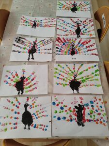 Na zdjęciu widać stół przykryty jasnym obrusem, na którym rozłożonych jest osiem prac plastycznych wykonanych przez dzieci. Każda praca przedstawia pawia – jego sylwetka została namalowana czarną farbą, a ogon ozdobiony kolorowymi kropkami.

Kropki tworzą wachlarz piór w żywych barwach: czerwonej, niebieskiej, zielonej, różowej, pomarańczowej i fioletowej. Widać, że dzieci używały techniki stemplowania palcami, ponieważ kropki mają różne rozmiary i kształty. Każdy paw wygląda nieco inaczej – różnią się układem kropek, intensywnością kolorów i kształtem sylwetki.

Całość sprawia wrażenie radosnej, kreatywnej wystawy prac przedszkolnych, pełnej kolorów i dziecięcej wyobraźni.