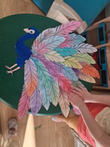 Na zdjęciu widać duży, kolorowy projekt plastyczny przedstawiający pawia. Jego ciało zostało wykonane z papieru i pomalowane na intensywny niebieski kolor, a ogon składa się z wielu papierowych piór w różnych barwach – różowych, fioletowych, niebieskich, zielonych, żółtych, pomarańczowych i czerwonych. Każde pióro jest osobno pokolorowane i wycięte, a następnie ułożone w wachlarz, tworząc efektowny, rozłożysty ogon.

Praca leży na okrągłym, zielonym stole. Po prawej stronie widać dziecko w pomarańczowych spodenkach i żółtej bluzce, które delikatnie dotyka piór, prawdopodobnie kończąc lub podziwiając swoją pracę. W tle widać fragmenty mebli i podłogi w sali przedszkolnej. Całość wygląda jak wspólna, kreatywna zabawa plastyczna.