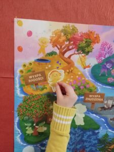 Na zdjęciu widać fragment kolorowego plakatu przedstawiającego bajkowe wyspy symbolizujące różne emocje. Na pierwszym planie znajduje się Wyspa Radości – pełna ciepłych barw, kwiatów, balonów i uśmiechniętych postaci. Widać tam żółte drzewo z kolorowymi liśćmi oraz postać wesołego stworka.

Dziecko w żółtym swetrze z białymi paskami na rękawie trzyma w dłoni papierową postać i przykleja ją lub przykłada do plakatu, dokładnie w miejscu oznaczonym jako „Wyspa Radości”. W tle widać fragment sąsiedniej wyspy – „Wyspy Znudzenia”, utrzymanej w ciemniejszych, szarych barwach.

Scena przedstawia moment zajęć edukacyjnych, prawdopodobnie dotyczących emocji i uczuć, w których dzieci uczą się rozpoznawać i nazywać różne stany emocjonalne.
