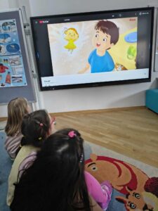 Na zdjęciu widać troje dzieci siedzących na kolorowym dywanie w sali przedszkolnej. Dzieci oglądają bajkę wyświetlaną na dużym ekranie dotykowym zawieszonym na ścianie. Na ekranie widać animowaną scenę przedstawiającą chłopca w niebieskiej koszulce oraz małą, żółtą wróżkę unoszącą się obok niego.

Dzieci siedzą blisko siebie, zwrócone w stronę ekranu, skupione na oglądaniu. W tle po lewej stronie znajduje się tablica z planszami edukacyjnymi, a po prawej fragment niebieskiej kanapy. Pomieszczenie jest jasne i uporządkowane, co sugeruje, że to sala przedszkolna lub szkolna przestrzeń do zajęć multimedialnych.



