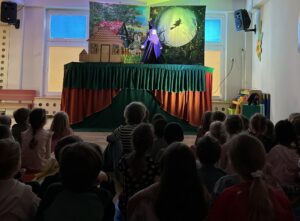 Na zdjęciu widać grupę dzieci siedzących na podłodze i oglądających teatrzyk kukiełkowy. Scena jest udekorowana kolorową tkaniną – dół w odcieniach zieleni i pomarańczu, a tło przedstawia domek w lesie oraz księżyc z lecącą czarownicą na miotle.

Na scenie znajduje się kukiełka czarownicy w długiej, fioletowej sukni i czarnym kapturze. Dzieci siedzą w skupieniu, zwrócone w stronę przedstawienia. W pomieszczeniu panuje półmrok, co podkreśla teatralny nastrój.

Całość wygląda jak spektakl dla przedszkolaków odbywający się w sali  przedszkolnej. Atmosfera jest pełna ciekawości i skupienia.
