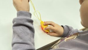 Na zdjęciu widać dziecko siedzące przy stole i trzymające w dłoniach żółtą ozdobę z filcu. Ozdoba ma przymocowany żółty sznurek, dzięki któremu można ją zawiesić, np. na gałązce lub choince. W dolnej części dekoracji widoczny jest różowy element, prawdopodobnie fragment ozdobnego wzoru lub wypełnienia. Dziecko ma na sobie szarą bluzę z nadrukiem postaci z kreskówki. Scena przedstawia moment tworzenia lub oglądania ręcznie wykonanej dekoracji, prawdopodobnie o tematyce świątecznej.