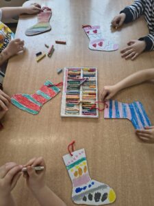 Na zdjęciu widać grupę dzieci siedzących przy stole i wykonujących pracę plastyczną. Na blacie leży kilka papierowych skarpetek, które dzieci kolorują kredkami świecowymi.

Każda skarpetka jest ozdobiona inaczej – widać wzory w paski, serduszka, kropki i fale, a także różne kolory: czerwony, niebieski, zielony, różowy, żółty i pomarańczowy. W środku stołu znajduje się pudełko z kredkami w wielu barwach, z którego dzieci korzystają podczas pracy.

Skarpetki mają przyczepione czerwone sznureczki, co sugeruje, że po zakończeniu zajęć będą mogły zostać powieszone jako dekoracja. Atmosfera zdjęcia jest radosna i twórcza – to prawdopodobnie zajęcia plastyczne z okazji Dnia Kolorowej Skarpetki.