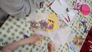 Na zdjęciu widać stół przykryty ceratą w zielono-białą kratkę z motywem wiśni, na którym odbywają się zajęcia plastyczne. Na stole rozłożone są serwetki z kwiatowymi wzorami – przedstawiają bukiety wiosennych kwiatów, takich jak żonkile, bratki i fiołki. Obok leżą nożyczki, pędzelki, klej w butelce z aplikatorem oraz białe tacki służące do pracy. Widać ręce dzieci i dorosłych, które wycinają lub przyklejają elementy z serwetek, prawdopodobnie tworząc ozdoby wielkanocne techniką decoupage. Na stole leżą też kolorowe kartoniki i wycięte fragmenty kwiatów, a w tle widać różowe jajko z papieru. Cała scena przedstawia twórcze, spokojne zajęcia plastyczne, pełne wiosennych kolorów i świątecznego nastroju.