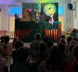 Na zdjęciu widać grupę dzieci siedzących na podłodze i oglądających teatrzyk kukiełkowy. Scena jest ustawiona na podwyższeniu, przykryta zielono-pomarańczoną tkaniną, a w tle znajdują się dwa kolorowe obrazy – po lewej stronie domek w lesie, a po prawej księżyc z lecącą czarownicą na miotle.

Na scenie stoją cztery kukiełki: po lewej czarownica w czarnej pelerynie i kapeluszu, obok niej postać w kolorowym stroju, dalej zwierzę przypominające wilka lub psa, a po prawej dziewczynka w różowej sukience i czapeczce.

Dzieci siedzą w skupieniu, zwrócone w stronę sceny. W pomieszczeniu panuje półmrok, a światło skierowane jest na teatrzyk, co tworzy magiczny, bajkowy nastrój. Całość wygląda jak przedstawienie dla przedszkolaków 