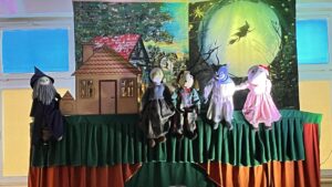 Na zdjęciu widać scenę teatrzyku kukiełkowego. Tło przedstawia nocny krajobraz – po lewej stronie znajduje się domek z czerwonym dachem wśród drzew, a po prawej duży, zielony księżyc, na tle którego widać czarownicę lecącą na miotle.

Na scenie ustawionej z zielono-pomarańczonej tkaniny znajduje się pięć kukiełek:

po lewej czarodziej z długą siwą brodą i spiczastym kapeluszem,
obok niego postać w niebieskiej sukni,
dalej postać w ciemnym stroju,
następnie niebieski wilk lub diabełek w jasnej koszuli,
po prawej dziewczynka w różowej sukience i czerwonej opasce.
Scena jest oświetlona kolorowym światłem, co nadaje przedstawieniu bajkowy i magiczny nastrój. Całość wygląda jak spektakl dla dzieci, inspirowany klasycznymi baśniami.