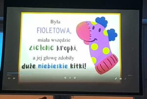 Na zdjęciu widać ekran, na którym wyświetlony jest slajd z kolorową ilustracją i tekstem. Po prawej stronie znajduje się rysunek fioletowej skarpetki z zielonymi kropkami, różowym noskiem i dużymi niebieskimi kitkami na górze. Skarpetka ma uśmiechniętą buzię i wygląda wesoło, jak postać z bajki dla dzieci.

Po lewej stronie widnieje tekst w różnych kolorach i czcionkach: fioletowej, zielonej, czarnej i niebieskiej. Całość jest utrzymana w radosnym, dziecięcym stylu – wygląda jak fragment prezentacji lub bajki czytanej podczas zajęć w przedszkolu.