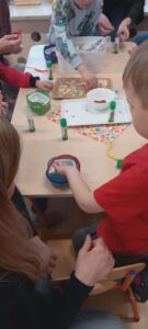 Na zdjęciu widać grupę dzieci siedzących przy stole i biorących udział w zajęciach plastycznych. Na stole leżą różne materiały: kleje w sztyfcie, kolorowe miseczki, tacki z drobnymi elementami dekoracyjnymi oraz pojemniki z watą lub masą plastyczną. Dzieci, ubrane w kolorowe koszulki, skupiają się na swoich pracach – jedno z nich w czerwonej koszulce trzyma mały pojemnik, do którego coś wkłada lub ugniata. Obok siedzi dorosła osoba, prawdopodobnie nauczycielka lub opiekunka, pomagająca dziecku. Na stole leży też kolorowa serwetka w kropki, a cała scena odbywa się w jasnej sali przedszkolnej. Atmosfera jest twórcza i radosna – dzieci wspólnie wykonują pracę plastyczną, prawdopodobnie związaną z wiosną lub Wielkanocą.