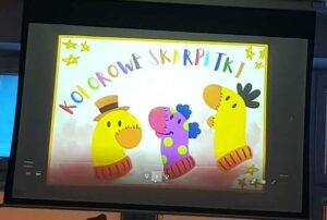 Na zdjęciu widać ekran, na którym wyświetlony jest kolorowy slajd z napisem „Kolorowe skarpetki”. Litery tytułu są w różnych barwach – czerwonej, fioletowej, niebieskiej, zielonej, pomarańczowej i żółtej – co nadaje całości radosny, dziecięcy charakter.

Pod napisem znajdują się trzy uśmiechnięte skarpetki przedstawione jak postacie z bajki.

Po lewej stronie jest żółta skarpetka w brązowym kapeluszu.
W środku znajduje się różowo-fioletowa skarpetka w zielone grochy, z niebieskimi kitkami.
Po prawej stronie widoczna jest kolejna żółta skarpetka z czarnym pióropuszem lub fryzurą z włóczki.
Tło jest jasne, ozdobione żółtymi gwiazdkami w rogach. Całość wygląda jak kadr z bajki lub prezentacji dla dzieci – wesoła, kolorowa i pełna pozytywnej energii.