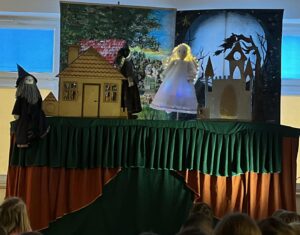 Na zdjęciu widać scenę teatrzyku kukiełkowego przedstawiającą baśniowy krajobraz. Na podwyższeniu przykrytym zieloną tkaniną ustawiono dekoracje: po lewej stronie stoi model domku z brązowym dachem i drzwiami, przypominający chatkę w lesie, a po prawej czerwony zamek z wieżyczkami i oknami, podświetlony od środka ciepłym światłem.

Pomiędzy nimi znajduje się kukiełka postaci w ciemnym stroju z kapturem, prawdopodobnie czarownicy lub bohaterki bajki. W tle rozciąga się malowany pejzaż nocnego lasu z dużym, pełnym księżycem, na tle którego widać czarownicę lecącą na miotle.

Całość tworzy nastrojową, magiczną scenę w klimacie klasycznej baśni – pełną kolorów, światła i tajemniczej atmosfery.