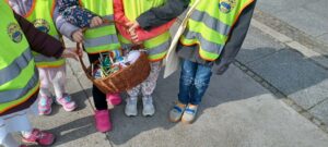 Na zdjęciu widać grupę dzieci ubranych w żółte kamizelki odblaskowe z emblematem przedszkola. Dzieci stoją na chodniku i wspólnie trzymają wiklinowy koszyk wypełniony kolorowymi ozdobami wielkanocnymi — widać w nim m.in. jajka, króliczki i wstążki. Ubrane są w ciepłe ubrania: kurtki, spodnie, legginsy i sportowe buty w różnych kolorach. Scena przedstawia moment podczas spaceru lub akcji wielkanocnej, w której dzieci niosą własnoręcznie przygotowane dekoracje.
