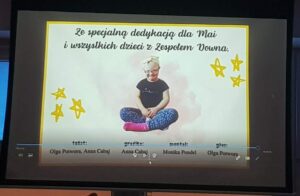 Na zdjęciu widać ekran, na którym wyświetlony jest slajd z dedykacją. W centralnej części znajduje się zdjęcie dziewczynki siedzącej po turecku na jasnym tle. Ma na sobie czarną koszulkę, niebieskie legginsy w wzory i różowe skarpetki. Na głowie ma opaskę z kokardką.

Nad zdjęciem widnieje napis:
„Ze specjalną dedykacją dla Mai i wszystkich dzieci z Zespołem Downa.”

Tekst jest zapisany ozdobną czcionką w kolorze czarnym, a całość otacza żółta ramka. Po bokach znajdują się narysowane żółte gwiazdki, które dodają pracy ciepłego, radosnego charakteru.

Na dole slajdu umieszczono podpisy osób zaangażowanych w projekt – przy słowach tekst, grafika, montaż i muzyka widnieją imiona i nazwiska twórców. Całość wygląda jak końcowy kadr filmu lub prezentacji przygotowanej z okazji Dnia Zespołu Downa.