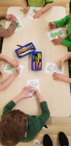 Na zdjęciu widać grupę dzieci siedzących przy prostokątnym stole i kolorujących obrazki. Każde dziecko ma przed sobą kartkę z tą samą ilustracją przedstawiającą małego zielonego stworka – prawdopodobnie postać z bajki lub zajęć tematycznych.

Na środku stołu stoją dwa pojemniki z kredkami ołówkowymi i pastelami w różnych kolorach. Dzieci skupione są na pracy, większość z nich używa zielonych kredek, co widać po kolorze postaci na rysunkach.

Dzieci ubrane są w odcienie zieleni i brązu, co może sugerować, że zajęcia mają motyw przewodni związany z kolorem zielonym lub przyrodą. Scena jest spokojna i pełna skupienia – widać, że dzieci z zaangażowaniem wykonują swoje prace plastyczne.