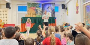 Na zdjęciu widać grupę dzieci siedzących na podłodze w sali przedszkolnej lub szkolnej, oglądających występ teatralny. Przed nimi stoi mężczyzna w jasnej koszulce i ciemnych spodniach, który prowadzi przedstawienie lub rozmawia z dziećmi. Obok niego stoi mała dziewczynka w różowej bluzce i pomarańczowej spódniczce, zwrócona w jego stronę.

W tle znajduje się scena teatrzyku kukiełkowego przykryta zielono-pomarańczoną tkaniną. Dekoracja przedstawia domek z czerwonym dachem po lewej stronie oraz nocne niebo z księżycem i czarownicą na miotle po prawej.

Dzieci siedzą w skupieniu, wiele z nich ma podniesione ręce, jakby chciały odpowiedzieć na pytanie lub wziąć udział w zabawie. Wnętrze sali jest jasne i kolorowe, na ścianach widać ozdoby w kształcie zwierząt. Atmosfera jest radosna i pełna zaangażowania.