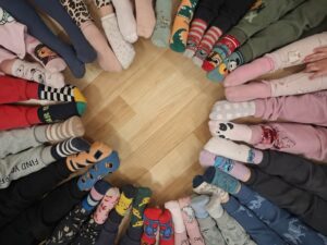 Na zdjęciu widać grupę osób siedzących w kręgu na podłodze, z nogami skierowanymi do środka. Każdy ma na sobie kolorowe, nie do pary skarpetki – w paski, kropki, wzory zwierzęce, z postaciami z bajek i różnymi motywami.

Podłoga jest drewniana, a w środku kręgu pozostaje puste miejsce w kształcie koła. Układ nóg tworzy efektowną, symetryczną kompozycję pełną barw i wzorów.

Zdjęcie symbolizuje Dzień Kolorowej Skarpetki, obchodzony 21 marca – dzień solidarności z osobami z zespołem Downa. Atmosfera fotografii jest radosna, przyjazna i pełna pozytywnej energii