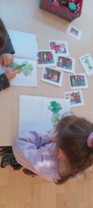 Na zdjęciu widać dwoje dzieci siedzących przy stole i kolorujących obrazki przedstawiające zielonego stworka w kapeluszu i płaszczu. Każde dziecko ma przed sobą zeszyt, w którym znajduje się ta sama ilustracja – postać jest już częściowo pokolorowana na zielono.

Na stole, obok zeszytów, leży kilka kolorowych zdjęć i karteczek – przedstawiają one grupy dzieci oraz tę samą postać, którą dzieci kolorują. W prawym górnym rogu stoi pojemnik z przyborami plastycznymi: kredkami, flamastrami i nożyczkami.

Scena wygląda na zajęcia plastyczne w przedszkolu lub szkole, podczas których dzieci pracują nad tematem związanym z zielonym bohaterem. Atmosfera jest spokojna i skupiona – dzieci z zaangażowaniem wykonują swoje rysunki.