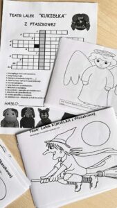 Na zdjęciu widać kilka czarno-białych kartek z materiałami edukacyjnymi dla dzieci, związanych z Teatrem Lalek „Kukiełka” z Ptaszkowej.

Na górze znajduje się krzyżówka z tytułem „Teatr Lalek ‘Kukiełka’ z Ptaszkowej”. Pod nią widoczne są rysunki lalek – m.in. czarownicy, anioła i misia. Poniżej krzyżówki umieszczono listę pytań oraz miejsce na wpisanie hasła.

Na pozostałych kartkach znajdują się kolorowanki przedstawiające postacie z przedstawień:

anioła z aureolą i skrzydłami,
czarownicę lecącą na miotle na tle księżyca.
Kartki leżą na drewnianym blacie, ułożone częściowo jedna na drugiej. Całość wygląda jak materiały plastyczno-edukacyjne przygotowane dla dzieci po spektaklu teatralnym.