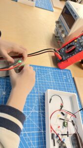 Na zdjęciu widać osobę, która pracuje przy stanowisku elektronicznym. Na niebieskiej macie technicznej leży płytka z podłączonymi przewodami, a osoba trzyma ją w dłoniach, sprawdzając działanie układu — na płytce świeci się zielona dioda LED. Obok znajduje się czerwony multimetr cyfrowy z podłączonymi przewodami pomiarowymi oraz zasilacz laboratoryjny. Na tacy leżą różne elementy elektroniczne: przewody, rezystory, diody i małe płytki. Scena przedstawia zajęcia z elektroniki, podczas których uczestnik testuje prosty obwód elektryczny.