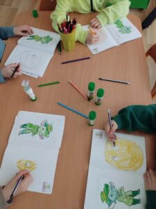 Na zdjęciu widać czworo dzieci siedzących przy drewnianym stole i wykonujących prace plastyczne. Każde dziecko ma przed sobą otwarty zeszyt lub blok rysunkowy, w którym koloruje tę samą postać – małego zielonego stworka w kapeluszu i płaszczu.

Na stole leży kilka kredek w różnych kolorach, głównie zielonych, żółtych i niebieskich, a także trzy kleje w sztyfcie. W środku stołu stoi zielony kubek wypełniony kredkami.

Dzieci są skupione na pracy – widać, że starannie kolorują swoje rysunki. Dominują odcienie zieleni, co nadaje całej scenie spójny, wiosenny i radosny charakter. Zdjęcie przedstawia spokojny moment zajęć plastycznych, prawdopodobnie w przedszkolu lub wczesnoszkolnej klasie.