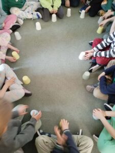 Na zdjęciu widać grupę dzieci siedzących w kręgu na podłodze, na jasnozielonym dywanie. Każde dziecko trzyma w rękach plastikowy kubeczek – biały, żółty lub jasnozielony. Część kubeczków stoi odwrócona dnem do góry przed dziećmi.

Dzieci siedzą blisko siebie, tworząc równy okrąg, i wyglądają na skupione. Wygląda na to, że biorą udział w zabawie rytmicznej lub integracyjnej, w której używa się kubeczków do wystukiwania rytmu lub przekazywania ich sobie w określonym tempie.

Scena ma spokojny, zorganizowany charakter i przedstawia moment wspólnej aktywności grupowej, prawdopodobnie w przedszkolu lub wczesnoszkolnej klasie.