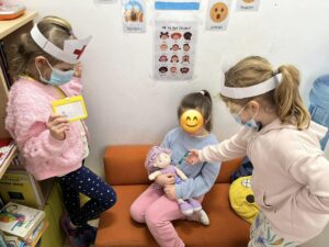 Na zdjęciu widać trzy dziewczynki bawiące się w lekarza w przedszkolu lub szkole. Dwie z nich mają na głowach papierowe opaski z czerwonym krzyżem, symbolizujące pielęgniarki lub lekarki, oraz maseczki ochronne. Jedna z dziewczynek trzyma w ręku małą tabliczkę, a druga „bada” koleżankę siedzącą na pomarańczowej kanapie.

Dziewczynka siedząca na kanapie trzyma na kolanach lalkę, która prawdopodobnie jest „pacjentem”. W tle widać ścianę z plakatami edukacyjnymi przedstawiającymi emocje i twarze dzieci. Scena pokazuje zabawę tematyczną, w której dzieci uczą się empatii i troski o innych poprzez odgrywanie ról.