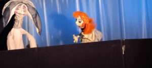 Na zdjęciu widzę scenę z przedstawienia teatru lalek. Oto szczegółowy opis:

Postacie:

Po lewej stronie: Lalka w jasnym, beżowym kostiumie z charakterystycznym długim, spiczastym kapturem lub czapką. Postać ma wyrazistą, białą twarz z malowanymi różowymi policzkami i wydaje się być w dramatycznej pozie - ręce uniesione do głowy w geście rozpaczy lub zaskoczenia.
Po prawej stronie: Lalka z jaskrawymi, pomarańczowymi włosami ułożonymi w charakterystyczną fryzurę. Ma duże, wyraziste oczy i ubrana jest w jasny kostium. Postać gestykuluje rękami, jakby była w trakcie rozmowy lub opowiadania.
Scenografia:

Tło stanowi gładka, niebieska kurtyna, która tworzy klasyczne teatralne tło
Na pierwszym planie widoczna jest czarna, matowa zasłona lub parawan, za którym ukrywają się lalkarze
Oświetlenie jest jasne i równomierne, skupiające uwagę na lalkach
Atmosfera:
Scena wygląda na moment z przedstawienia dla dzieci - lalki są kolorowe, wyraziste i pełne charakteru. Ich pozy sugerują dynamiczną interakcję między postaciami.

Czy chciałbyś, żebym pomógł Ci stworzyć coś związanego z tym zdjęciem? Na przykład plakat teatralny, zaproszenie na przedstawienie lub inny projekt graficzny?