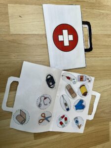 Na zdjęciu widać papierową pracę plastyczną przedstawiającą apteczkę pierwszej pomocy. Apteczka została wykonana z białego papieru i ma narysowany uchwyt, dzięki czemu przypomina prawdziwą torbę medyczną. Na jej zewnętrznej stronie widnieje czerwone koło z białym krzyżem pośrodku — symbol pierwszej pomocy.

Wewnątrz apteczki znajdują się narysowane i wycięte obrazki przedstawiające różne przedmioty medyczne: bandaż, nożyczki, stetoskop, ciśnieniomierz, termometr, plaster, rękawiczki, tabletki, tubkę z maścią oraz strzykawkę. Każdy element jest umieszczony w osobnym okręgu, co nadaje pracy porządek i przejrzystość.

Całość leży na drewnianej podłodze. Praca wygląda jak ciekawy projekt edukacyjny dla dzieci, pomagający im poznać wyposażenie apteczki i zasady udzielania pierwszej pomocy.