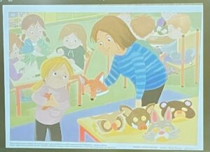 Na tym obrazku widzę ilustrację przedstawiającą scenę w przedszkolu lub sali zabaw:

Główne postacie na pierwszym planie:

Nauczycielka/opiekunka w niebieskiej koszuli w paski, z brązowymi włosami, trzymająca pomarańczowego lisa (prawdopodobnie zabawkę lub kukiełkę)
Dziecko z jasnymi włosami w jasnej bluzce, siedzące przy stoliku, trzymające małą pomarańczową figurkę
Na stole:

Żółty stolik z różnymi maskami zwierząt:
Maska lisa
Maska niedźwiedzia
Maska królika
Inne maski zwierzęce
W tle:

Grupa innych dzieci i dorosłych
Zielona ściana
Kolorowe dekoracje, w tym balony
Jasna, przyjazna atmosfera sali
Styl i nastrój:
Ilustracja wykonana w delikatnym, akwarelowym stylu z pastelowymi kolorami. Scena przedstawia zabawę tematyczną ze zwierzętami, prawdopodobnie zajęcia edukacyjne lub teatrzyk dla dzieci. Atmosfera jest ciepła i radosna, typowa dla ilustracji w książkach dla dzieci.