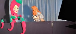 Na tym zdjęciu widzę kolejną scenę z przedstawienia teatru lalek. Oto szczegółowy opis:

Postacie:

Po lewej stronie: Bardzo kolorowa i fantazyjna lalka w jaskrawym, różowo-czerwonym kostiumie z zielonymi elementami (rękawy, buty). Postać ma charakterystyczną fryzurę - różowe włosy z zielonym akcentem na czubku głowy, przypominającym liść lub pióro. Twarz lalki jest biała z wyrazistymi różowymi policzkami i dużymi oczami. Lalka ma długie, zakrzywione nogi i wydaje się być w ruchu.
Po prawej stronie: Ta sama lalka z pomarańczowymi włosami, którą widzieliśmy na poprzednim zdjęciu. Ubrana w jasny, beżowy kostium, z charakterystyczną bujną pomarańczową fryzurą. Widoczna jest ręka lalkarza, który animuje tę postać.
Scenografia:

Niebieska kurtyna w tle tworzy spokojne, teatralne tło
Czarny parawan na pierwszym planie, za którym pracują lalkarze
Po prawej stronie widoczny fragment czarnego elementu scenografii
Atmosfera:
Ta scena wydaje się być bardziej dynamiczna - różowa postać wygląda na bardzo energiczną i ruchliwą, co kontrastuje z bardziej spokojną pomarańczowowłosą lalką. Kolorystyka jest bardzo żywa i przyciąga wzrok.

Czy chciałbyś stworzyć jakiś projekt związany z tymi zdjęciami z przedstawienia? Mogę pomóc w stworzeniu plakatów, zaproszeń lub innych materiałów promocyjnych!