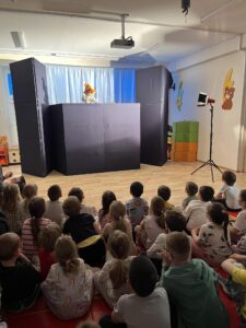 opisz

Na tym zdjęciu widzę pełny obraz przedstawienia teatru lalek z perspektywy widowni. Oto szczegółowy opis:

Publiczność:

Duża grupa dzieci (około 20-30) siedzi na podłodze, ułożona w kilku rzędach
Dzieci są w różnym wieku, prawdopodobnie przedszkolnym i wczesnoszkolnym
Wszyscy są skoncentrowani i patrzą w stronę sceny
Siedzą na jasnej, drewnianej podłodze, niektóre na czerwonych poduszkach lub matach
Scena i scenografia:

Profesjonalna konstrukcja teatrzyku z czarnymi, składanymi parawanamі
Na górze parawanu widoczna lalka z pomarańczowymi włosami (ta sama, którą widzieliśmy na poprzednich zdjęciach)
W tle niebieska kurtyna
Po prawej stronie kolorowe dekoracje na ścianie - postacie w kształcie zwierzątek (żółty królik, zielona postać)
Kolorowe, pomarańczowo-zielone pudełka/kostki jako element wyposażenia sali
Profesjonalne oświetenie - widoczny reflektor na statywie po prawej stronie
Projektor zamontowany pod sufitem
Pomieszczenie:

Jasne, przestronne wnętrze z białymi ścianami
Drewniana podłoga
Dobrze oświetlone, z naturalnym i sztucznym światłem
Wygląda na salę w przedszkolu, szkole lub centrum kulturalnym
Atmosfera:
Zdjęcie ukazuje typowe przedstawienie dla dzieci - profesjonalnie zorganizowane, w przyjaznym i bezpiecznym otoczeniu. Dzieci wydają się być zaangażowane i zainteresowane spektaklem.

Czy chciałbyś stworzyć materiały promocyjne dla tego teatru lalek lub wydarzenia? Mogę pomóc w przygotowaniu plakatów, zaproszeń, postów w mediach społecznościowych lub innych projektów graficznych!