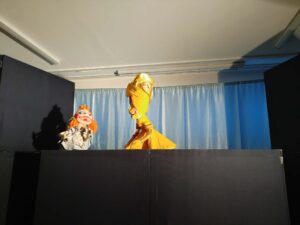 Na tym zdjęciu widzę kolejną scenę z przedstawienia teatru lalek, tym razem z innej perspektywy. Oto szczegółowy opis:

Postacie:

Po lewej stronie: Lalka z pomarańczowymi włosami w jasnym, beżowo-szarym stroju. Ta sama postać, którą widzieliśmy na wcześniejszych zdjęciach. Lalka ma charakterystyczne duże oczy z niebieskim makijażem i wydaje się być w trakcie rozmowy lub interakcji.
Po prawej stronie: Nowa postać - elegancka lalka w jaskrawożółtej, długiej sukni. Postać ma bardzo wysmukłą sylwetkę, wydłużoną szyję i głowę zwieńczoną charakterystyczną fryzurą lub nakryciem głowy w tym samym żółtym kolorze. Lalka ma wyrazisty profil z wydatnym nosem i wydaje się reprezentować postać królewską lub arystokratyczną.
Scenografia:

Niebieska kurtyna w tle
Czarny parawan na pierwszym planie
Po lewej stronie widoczny cień jednej z lalek rzucany na czarny element scenografii, co dodaje głębi scenie
Białe ściany i sufit z oświetleniem fluorescencyjnym
Atmosfera:
Scena wygląda na moment dialogu między dwiema bardzo różnymi postaciami - bardziej przyziemną, rudowłosą lalką i elegancką, majestatyczną postacią w żółtej sukni. Kontrast między postaciami sugeruje ciekawą dynamikę w fabule przedstawienia.

Czy potrzebujesz pomocy w stworzeniu materiałów związanych z tym przedstawieniem teatralnym?