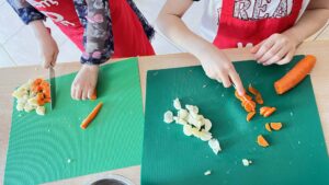 Na zdjęciu widać dwoje dzieci krojących warzywa na zielonych matach kuchennych. Na matach leżą kawałki marchewki i pietruszki, a obok widać jeszcze niepokrojoną marchewkę. Dzieci używają noży, trzymając warzywa ostrożnie, a każde z nich ma na sobie czerwony fartuszek ochronny.

Scena przedstawia moment zajęć kulinarnych, podczas których dzieci uczą się krojenia warzyw i przygotowywania posiłku. Atmosfera jest spokojna i skupiona, a praca odbywa się w czystym, dobrze oświetlonym pomieszczeniu.