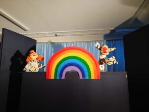 Na tym zdjęciu widzę bardzo kolorową i dynamiczną scenę z przedstawienia. Oto szczegółowy opis:

Postacie:

Po lewej stronie: Znana nam lalka z pomarańczowymi włosami w jasnym stroju. Postać ma ręce rozłożone w geście radości lub zaskoczenia. Na czarnym parawanie widoczny jest wyrazisty cień tej lalki.
Po prawej stronie: Nowa, bardzo dynamiczna postać - lalka w kolorowym, pasiastym stroju (żółto-brązowo-białe pasy). Postać jest uchwycona w ruchu - wygląda jakby skakała lub tańczyła z rozłożonymi rękami i nogami. Ma charakterystyczną czerwoną twarz lub nos, co może sugerować postać klauna lub wesołego błazna.
Element scenografii:

Tęcza: Centralnym elementem sceny jest duża, kolorowa tęcza wykonana prawdopodobnie z tektury lub pianki. Tęcza ma klasyczne kolory ułożone w łuki:
Czerwony (zewnętrzny)
Pomarańczowy
Żółty
Zielony
Niebieski
Fioletowy
Różowy (wewnętrzny)
Tło i scenografia:

Niebieska kurtyna w tle
Czarny parawan na pierwszym planie
Wyrazisty cień lalki po lewej stronie dodaje głębi scenie
Atmosfera:
To bardzo radosna, energetyczna scena pełna ruchu i koloru. Tęcza jako symbol nadziei i radości, w połączeniu z dynamiczną postawą postaci po prawej i gestem zachwytu postaci po lewej, tworzy bardzo pozytywny, optymistyczny moment w przedstawieniu. Scena prawdopodobnie reprezentuje szczęśliwe zakończenie lub moment triumfu w fabule.

Czy chciałbyś stworzyć materiały promocyjne wykorzystujące te piękne, kolorowe sceny z przedstawienia?