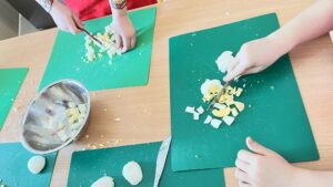 Na zdjęciu widać dzieci krojące ugotowane jajka na zielonych matach kuchennych. Każde dziecko ma przed sobą deskę i nóż, a na stole leży kilka już obranych jajek. Pokrojone kawałki jajek są odkładane do metalowej miski stojącej z boku.

Dzieci pracują ostrożnie, skupiając się na krojeniu. Ubrane są w czerwone fartuszki ochronne. Scena przedstawia zajęcia kulinarne, podczas których dzieci uczą się przygotowywania potrawy, prawdopodobnie sałatki z jajkiem.