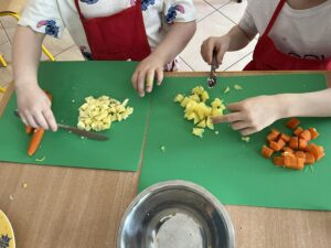 Na zdjęciu widać dwoje dzieci krojących warzywa na zielonych matach kuchennych. Dzieci mają na sobie czerwone fartuszki ochronne i białe koszulki z kolorowym nadrukiem. Na lewej macie leżą pokrojone kawałki ziemniaka oraz marchewka, a na prawej – kostki ugotowanego ziemniaka i marchewki.

Na środku stołu stoi metalowa miska, do której prawdopodobnie będą odkładane pokrojone składniki. Scena przedstawia zajęcia kulinarne, podczas których dzieci uczą się krojenia warzyw i przygotowywania potrawy, prawdopodobnie sałatki jarzynowej.