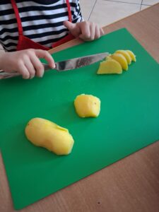 Na zdjęciu widać dziecko krojące ugotowanego ziemniaka na zielonej macie kuchennej. Dziecko trzyma nóż i ostrożnie tnie ziemniaka na równe plasterki. Obok na macie leży jeszcze jeden, niepokrojony ziemniak.

Dziecko ma na sobie koszulkę w czarno-białe paski oraz czerwony fartuszek ochronny. Scena przedstawia moment zajęć kulinarnych, podczas których dziecko uczy się bezpiecznego krojenia i przygotowywania składników do potrawy.