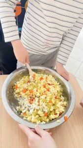 Na zdjęciu widać dziecko w bluzce w biało-granatowe paski, które miesza dużą metalową miskę pełną sałatki jarzynowej. W sałatce widać pokrojone składniki — ziemniaki, marchewkę, ogórki kiszone, jajka oraz groszek, połączone z majonezem.

Miska stoi na drewnianym stole, a drugie dziecko lub dorosły przytrzymuje ją z drugiej strony, pomagając w mieszaniu. Scena przedstawia moment wspólnego przygotowywania sałatki, prawdopodobnie podczas zajęć kulinarnych lub wspólnego gotowania.