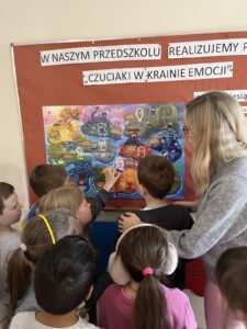Na tym zdjęciu widzę scenę z przedszkola lub szkoły podstawowej w Polsce:

Główne elementy:

Tablica informacyjna:

Czerwona tablica korkowa z napisami:
"W NASZYM PRZEDSZKOLU" i "REALIZUJEMY P..."
"CZUCIAKI W KRAINIE EMOCJI"
Plansza edukacyjna:

Kolorowa mapa/plansza przedstawiająca "Krainę Emocji" w stylu filmu "W głowie się nie mieści" (Inside Out)
Podzielona na różne strefy emocji z charakterystycznymi kolorami:
Żółta strefa (radość) - góra po lewej
Różowa/fioletowa strefa - lewa górna część
Niebieska strefa (smutek) - prawa górna część
Czerwona strefa (gniew) - centrum
Pomarańczowa strefa - dół
Ciemnoniebieska strefa (strach) - prawa strona
Na planszy widoczne są różne "wyspy" z napisami (np. "WYSPA RADOŚCI", "WYSPA SMUTKU" itp.)
Kolorowe postacie reprezentujące różne emocje
Osoby:

Nauczycielka/opiekunka po prawej stronie w szarej bluzie z jasnymi włosami
Grupa dzieci (około 8-10 dzieci) siedzących i stojących przed tablicą
Jedno dziecko wskazuje na planszę
Dzieci w różnym wieku, w casualowych ubraniach
Kontekst:
To zajęcia edukacyjne dotyczące rozpoznawania i nazywania emocji - program "Czuciaki w Krainie Emocji", który pomaga dzieciom w rozwoju inteligencji emocjonalnej. Dzieci uczą się identyfikować różne uczucia i emocje poprzez interaktywną mapę.