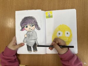 Na tym zdjęciu widzę dziecięcą pracę plastyczną w zeszycie:

Lewa strona zeszytu:

Rysunek postaci z fioletowymi włosami w stylu anime/manga
Postać ma:
Duże, niebiesko-zielone oczy
Różowe policzki
Uśmiech
Szarą bluzę/kurtkę z kieszenią
Szarą spódniczkę
Ciemne spodnie/legginsy
Bose stopy
Rysunek wykonany kredkami/flamastrami z kolorowaniem
Prawa strona zeszytu:

Duża żółta twarz/emotka narysowana żółtym flamastrem
Postać ma:
Wielkie, okrągłe białe oczy z ciemnymi źrenicami
Wyraz zaskoczenia lub strachu
Żółte włosy/kontury głowy wykonane grubymi kreskami
Dodatkowe elementy:

Mała naklejka w górnej części prawej strony przedstawiająca postać w szarym ubraniu na żółto-zielonym tle
Ręce dziecka w różowej bluzie trzymające zeszyt i czarny flamaster/marker z napisem "coloured p..." (prawdopodobnie "coloured pencil")
Drewniany stół/blat w tle
Kontekst:
Dziecko pracuje nad rysunkami postaci, prawdopodobnie inspirowanych filmem animowanym "W głowie się nie mieści" (Inside Out) - lewa postać może reprezentować smutek, a prawa radość. To kontynuacja zajęć o emocjach widocznych na poprzednich zdjęciach.