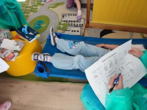 Na zdjęciu widać scenkę zabawy w lekarza zorganizowaną w przedszkolu. Na niebieskim leżaku leży dziecko, które jest „pacjentem”. Ma na sobie jasne spodnie z kolorowym nadrukiem i buty z motywem bajkowym. Obok, na żółtym stołku, leżą różne akcesoria medyczne — pudełka po plastrach, bandaże, rękawiczki i nożyczki.

Drugie dziecko, ubrane w zielony fartuch ochronny, siedzi obok i wypełnia kartę pacjenta, rysując lub zaznaczając odpowiednie pola na kartce. W tle widać fragment dywanu z kolorowym wzorem drogi i budynków. Scena przedstawia zabawę edukacyjną, w której dzieci uczą się roli lekarza i pacjenta w przyjazny, zabawowy sposób.