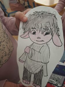 Na tym zdjęciu widzę dziecięcy rysunek trzymany w rękach:

Rysunek przedstawia:

Postać dziecka w stylu anime/manga z charakterystycznymi cechami:
Szare, potargane włosy z grzywką
Duże, okrągłe oczy z ciemnymi źrenicami
Różowe policzki i różowe uszy (przypominające uszy królika lub owcy)
Uśmiech z małym noskiem
Szara bluza/kurtka z kieszenią
Różowa spódniczka lub falbanka
Ciemne spodnie (szare lub czarne)
Bose stopy z zaznaczonymi palcami
Technika wykonania:

Rysunek wykonany czarnym flamastrem/pisakiem (kontury)
Kolorowanie kredkami lub ołówkami (szary, różowy)
Wyraźne kreskowanie przy wypełnianiu kolorami
Styl dziecięcy, ale staranny i przemyślany
Kontekst:

Ręce dziecka w różowej bluzie trzymają kartkę
W tle widoczne inne dzieci przy stolikach w sali przedszkolnej/szkolnej
Po prawej stronie widoczny fragment książki lub zeszytu z napisem "GO!!!"
To prawdopodobnie ta sama postać (Smutek z "W głowie się nie mieści"), którą widzieliśmy na poprzednim zdjęciu w zeszycie
Rysunek jest częścią zajęć edukacyjnych o emocjach prowadzonych w przedszkolu.