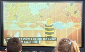 Na zdjęciu widzę scenę edukacyjną z projekcją multimedialną:

Główny element - ekran/projekcja:

Duży ekran telewizyjny/monitor wyświetlający animację edukacyjną
Kolorowa, dziecięca grafika z pastelowymi kolorami (miętowy, żółty, pomarańczowy, beżowy)
Treść na ekranie:

Postać pszczoły - duża, kreskówkowa pszczoła w czarno-żółte paski z przezroczystymi skrzydłami
Tło przedstawia plaster miodu (sześciokątne komórki w odcieniach żółci i pomarańżu)
Dolna część pokazuje strukturę plastra z geometrycznymi kształtami
Napisy w języku polskim: "Wn... zsz sobie a do tego ma ciężką... ta około 3000"
Tekst prawdopodobnie dotyczy informacji o pszczołach (waga, liczebność rodziny pszczelej)
Widzowie:

Dwoje dzieci siedzących przed ekranem
Widoczne z tyłu - brązowe włosy
Dzieci skupione na oglądaniu prezentacji
Jedno dziecko ma jasną kokardkę/spinką we włosach
Otoczenie:

Ciemniejsze pomieszczenie (prawdopodobnie sala lekcyjna lub świetlica)
Profesjonalna prezentacja multimedialna
Atmosfera:
Zdjęcie przedstawia typową sytuację edukacyjną - dzieci oglądają film lub prezentację edukacyjną o pszczołach. Kolorowa, przyjazna grafika i napisy w języku polskim wskazują na materiał dydaktyczny dostosowany do młodszych dzieci. To moment nauki przez zabawę - poznawanie świata przyrody, życia pszczół i ich znaczenia w ekosystemie w atrakcyjnej, wizualnej formie.